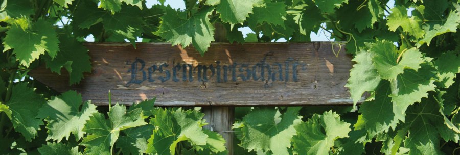 A wooden sign with the inscription 'Besenwirtschaft' is surrounded by green vine leaves., © SMG A wooden sign with the inscription 'Besenwirtschaft' is surrounded by green vine leaves., © SMG