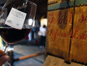 Ein Glas Rotwein mit dem Logo des Weinbaumuseums Stuttgart, im Hintergrund eine alte Holzpresse mit der Inschrift 'Gelobt sei Jesus Christus'., © Stuttgart-Marketing GmbH