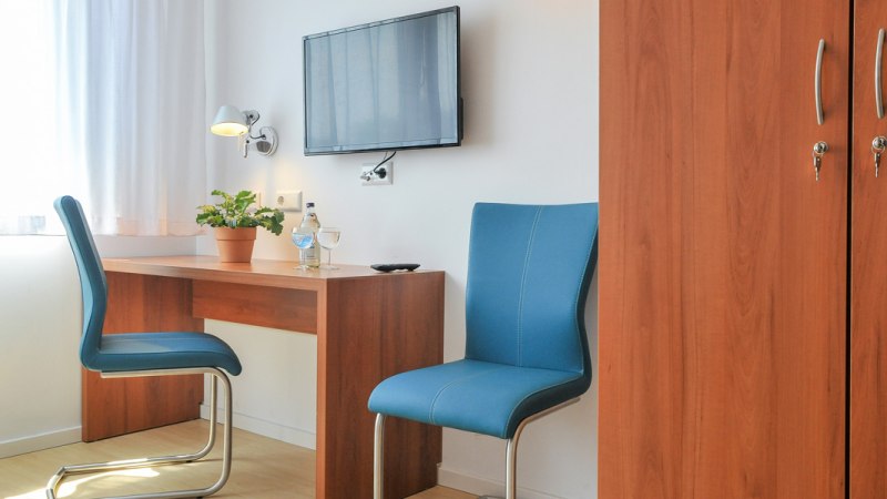Ein schlichtes Hotelzimmer mit Schreibtisch, zwei blauen St&uuml;hlen, einem Fernseher an der Wand und einem Kleiderschrank., &copy; G&auml;stehaus ABZ Stuttgart