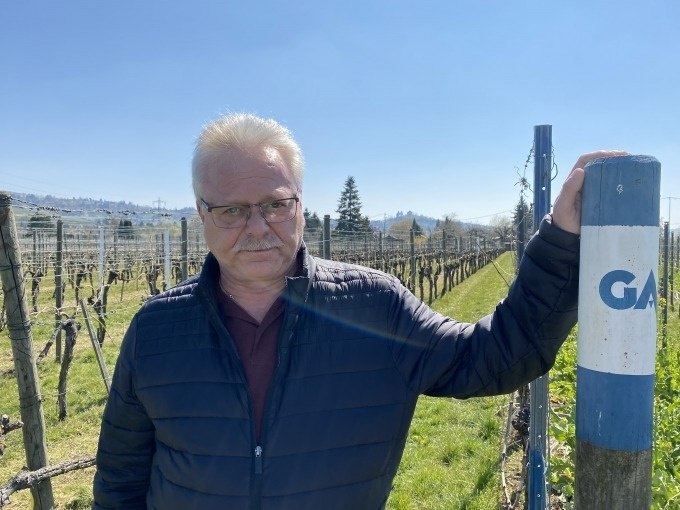 Ein Mann mit Brille steht in einem Weinberg und h&auml;lt einen Pfosten mit der Aufschrift 'GA'. Im Hintergrund sind Reben und blauer Himmel zu sehen., &copy; Weingenuss Stuttgart