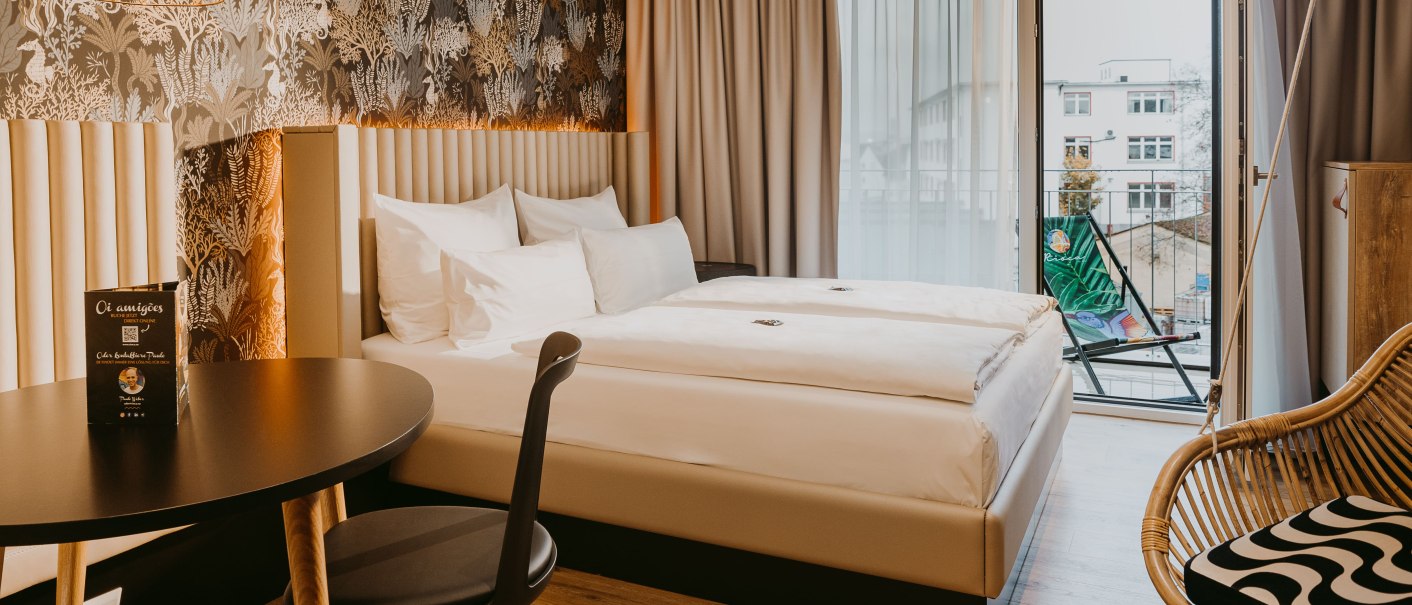Modern hotel room with double bed, stylish wallpaper, table and chair. Access to a balcony with deck chair. Bright, inviting atmosphere., © Rioca Stuttgart Posto 4 GmbH Modern hotel room with double bed, stylish wallpaper, table and chair. Access to a balcony with deck chair. Bright, inviting atmosphere., © Rioca Stuttgart Posto 4 GmbH