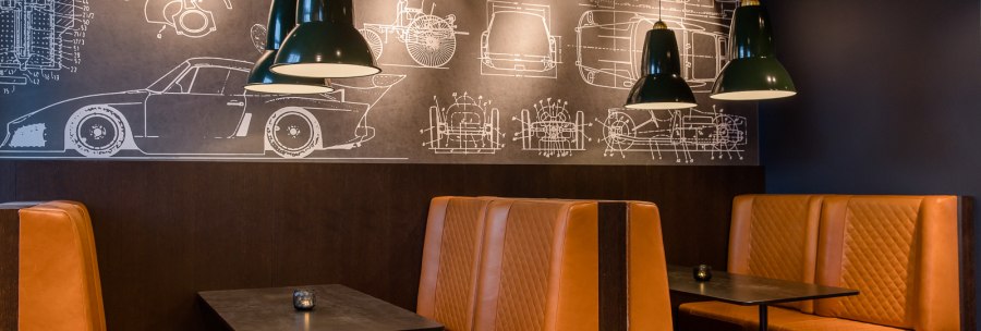 Restaurant mit braunen Ledersitzbänken, schwarzen Tischen und grünen Lampen. An der Wand sind technische Zeichnungen von Autos zu sehen., © TOMAS Restaurant mit braunen Ledersitzbänken, schwarzen Tischen und grünen Lampen. An der Wand sind technische Zeichnungen von Autos zu sehen., © TOMAS