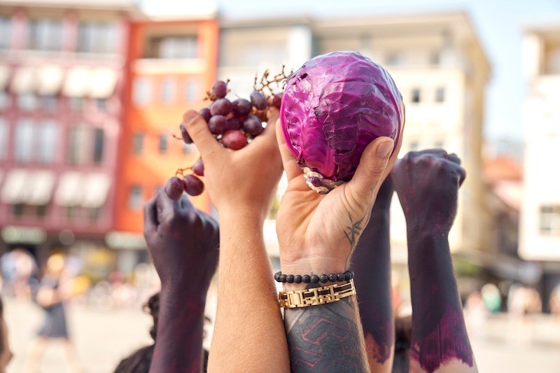 H&auml;nde halten roten Kohl und Trauben in die Luft. Die Arme sind teilweise bemalt. Im Hintergrund sind unscharfe Geb&auml;ude zu sehen., &copy; Theater Rampe