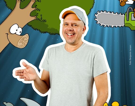 Ein Mann in einem grauen T-Shirt steht lächelnd vor einer Collage aus Cartoon-Tieren und -Objekten, darunter ein Nashorn, ein Baum und ein Fischglas., © Theaterhaus Stuttgart e.V. Ein Mann in einem grauen T-Shirt steht lächelnd vor einer Collage aus Cartoon-Tieren und -Objekten, darunter ein Nashorn, ein Baum und ein Fischglas., © Theaterhaus Stuttgart e.V.