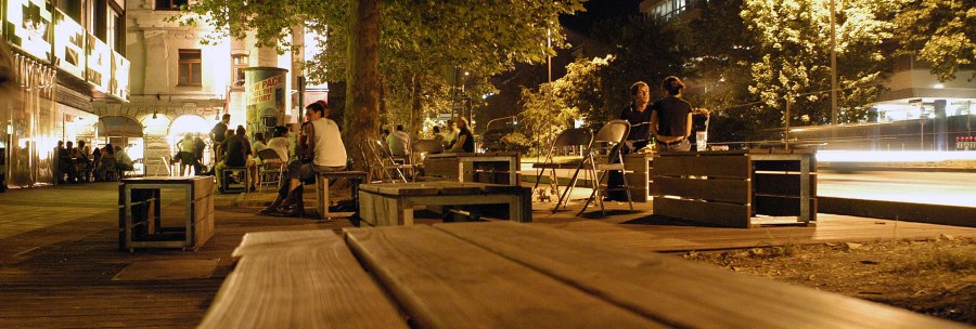 Nachtaufnahme der Theodor-Heuss-Straße in Stuttgart. Menschen sitzen auf Holzbänken unter Bäumen, umgeben von beleuchteten Gebäuden und Straßenlaternen., © Stuttgart-Marketing GmbH, Marc Gilardone Nachtaufnahme der Theodor-Heuss-Straße in Stuttgart. Menschen sitzen auf Holzbänken unter Bäumen, umgeben von beleuchteten Gebäuden und Straßenlaternen., © Stuttgart-Marketing GmbH, Marc Gilardone