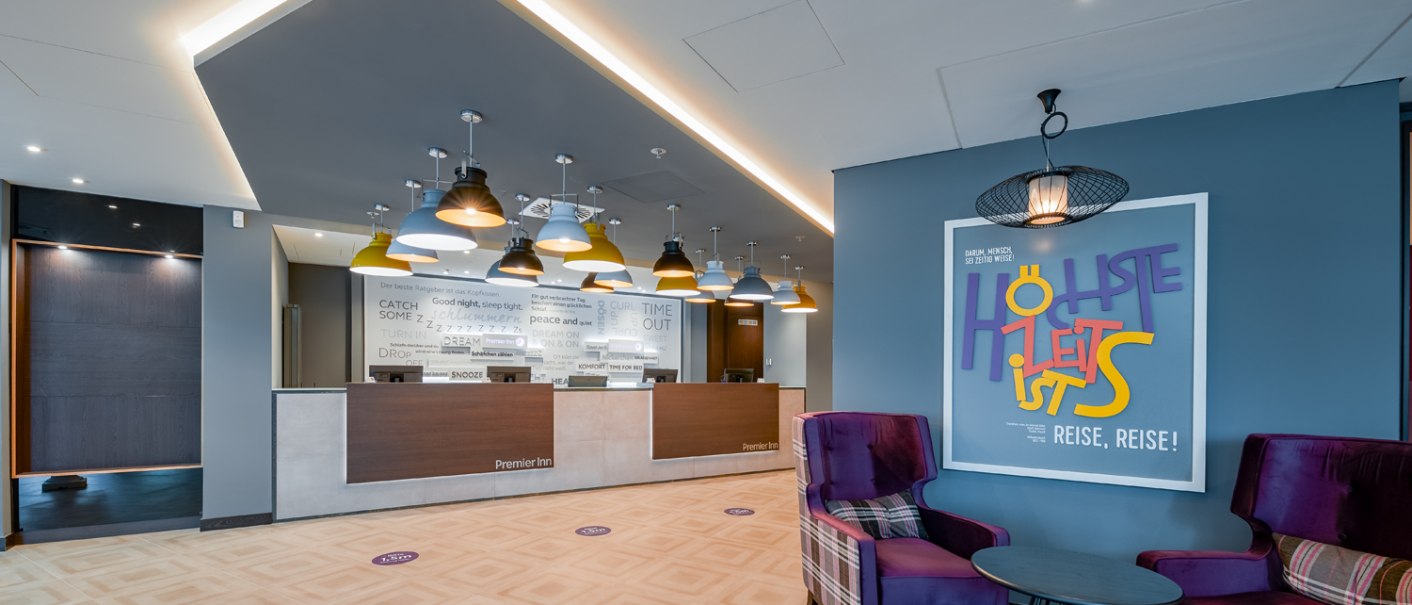 Moderne Hotelrezeption mit stilvollen Hängelampen, einem Sitzbereich mit lila Sesseln und einem auffälligen Wandplakat. Elegantes, einladendes Ambiente., © Premier Inn Moderne Hotelrezeption mit stilvollen Hängelampen, einem Sitzbereich mit lila Sesseln und einem auffälligen Wandplakat. Elegantes, einladendes Ambiente., © Premier Inn