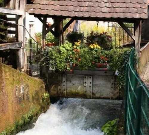 Ein altes Mühlrad mit Blumen geschmückt, umgeben von fließendem Wasser und einem Holzdach., © TOMAS