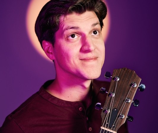 A man holds a guitar and looks upwards. The background is purple with a circle of light behind his head, which resembles a halo., &copy; Renitenztheater Stuttgart e.V.