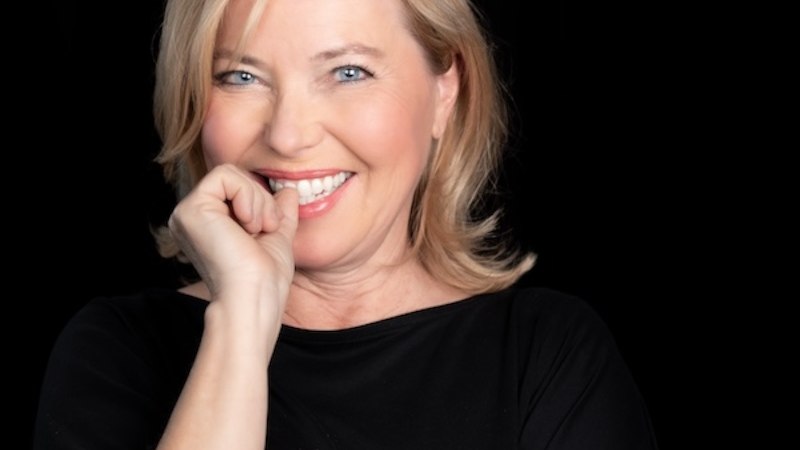 A woman with blonde hair smiles in a friendly manner and holds a hand in front of her mouth. She is wearing a black top against a black background., &copy; Renitenztheater Stuttgart e.V.
