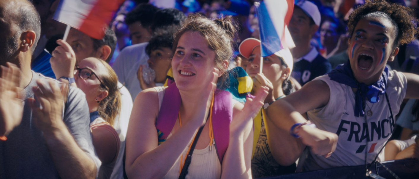 Menschen jubeln in einer Menge, einige halten französische Flaggen. Eine Frau trägt Gesichtsbemalung in den Farben der französischen Flagge., © Comme des Cinémas, Cinq de Trèfle Productions