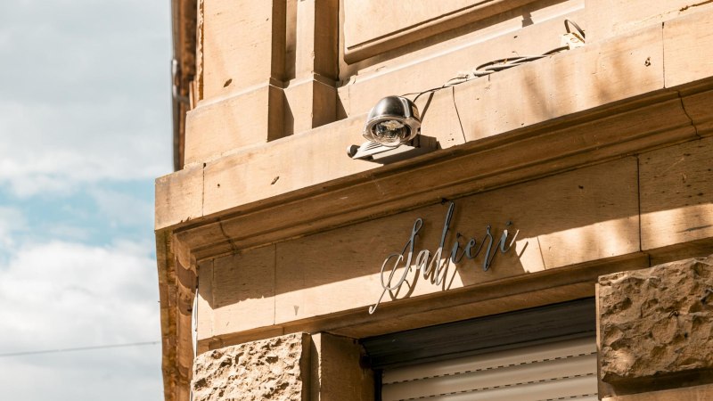 Fassade eines Gebäudes mit dem Schriftzug 'Salieri' und einer Überwachungskamera. Die Sonne wirft Schatten auf die sandfarbene Wand., © SMG, Sarah Schmid Fassade eines Gebäudes mit dem Schriftzug 'Salieri' und einer Überwachungskamera. Die Sonne wirft Schatten auf die sandfarbene Wand., © SMG, Sarah Schmid