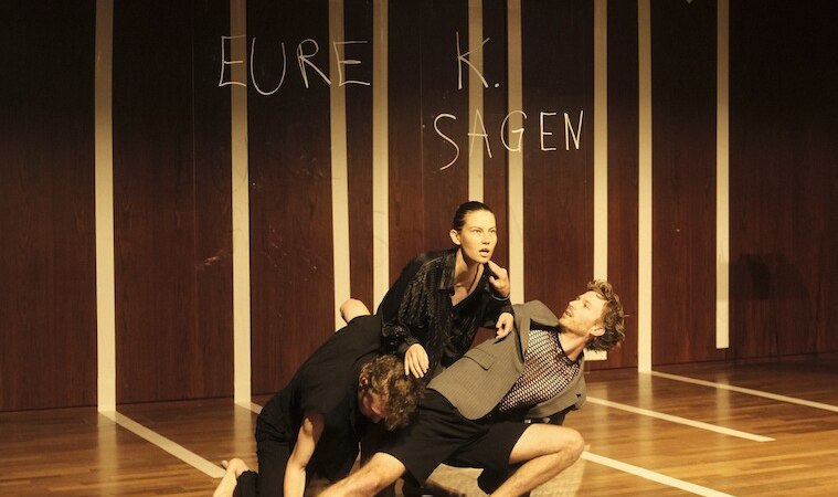 Three people in a dynamic dance pose on a stage. 'PADUM' is written in the background. The scene is artistic and expressive., &copy; Wilhelma Theater