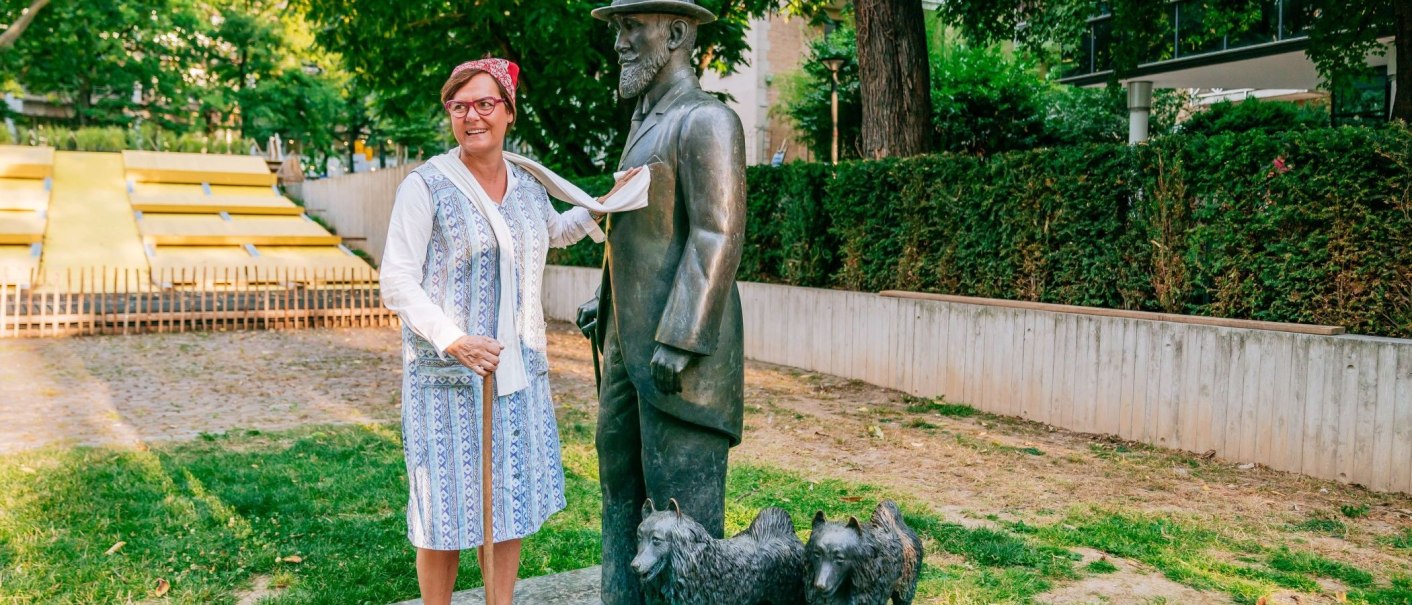 Frau in traditioneller Kleidung posiert lächelnd neben einer Bronzestatue eines Mannes mit Hut und zwei Hunden im Freien., © Thomas Niedermüller
