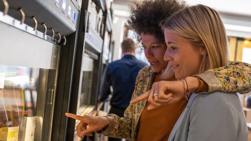 Zwei Frauen stehen vor einem Weinspender und zeigen auf verschiedene Weinflaschen. Im Hintergrund sind weitere Flaschen und Gläser zu sehen., © Stuttgart Marketing GmbH, Fotografin Martina Denker