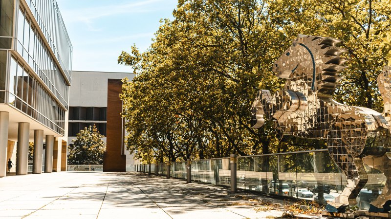 Moderne Glasfassade neben einer glänzenden Pferdeskulptur aus Spiegeln, umgeben von Bäumen und blauem Himmel., © Sarah Schmid