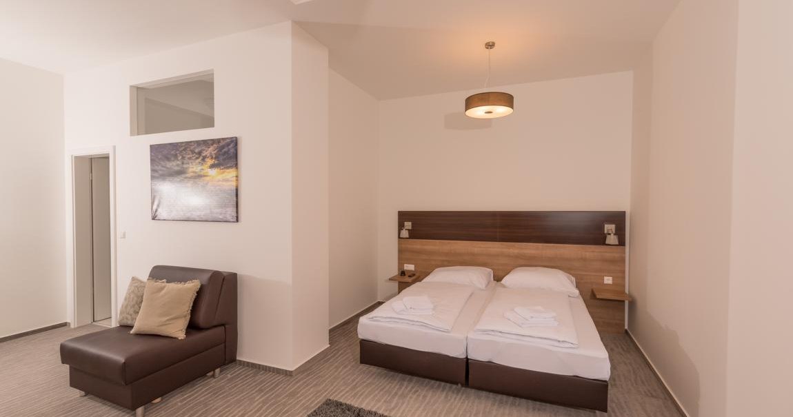 Ein modernes Hotelzimmer mit einem Doppelbett, einem braunen Sofa und einem Wandbild. Der Raum ist in neutralen Tönen gehalten., © TOMAS