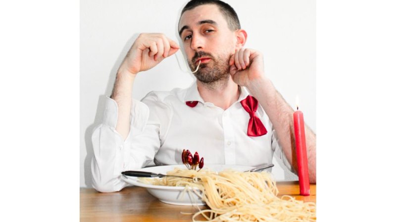 Ein Mann in weißem Hemd mit roter Fliege isst lässig Spaghetti. Eine rote Kerze steht auf dem Tisch und sorgt für eine humorvolle Atmosphäre., © Fotocredit Theater La Lune Stuttgart Ein Mann in weißem Hemd mit roter Fliege isst lässig Spaghetti. Eine rote Kerze steht auf dem Tisch und sorgt für eine humorvolle Atmosphäre., © Fotocredit Theater La Lune Stuttgart