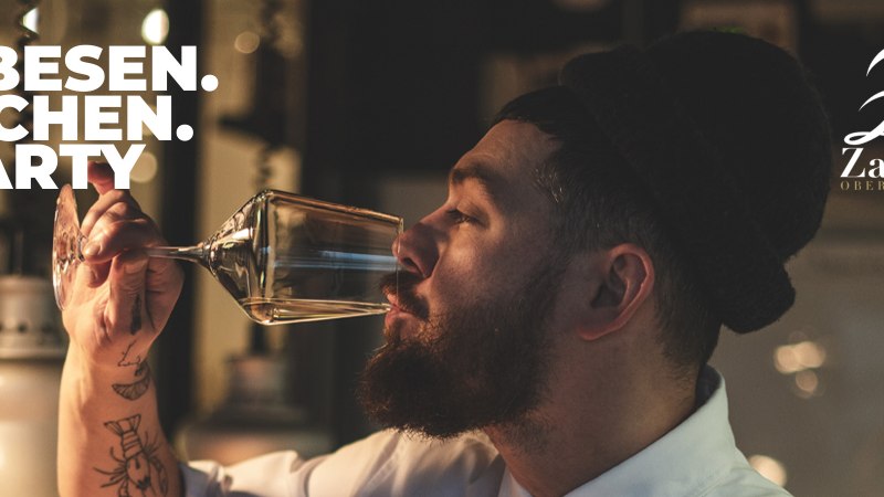 Ein Mann mit Bart genie&szlig;t ein Glas Wein. Im Hintergrund das Logo des Weinguts Zai&szlig;. Text: Besen-K&uuml;chen-Party., &copy; Weingut Zai&szlig;