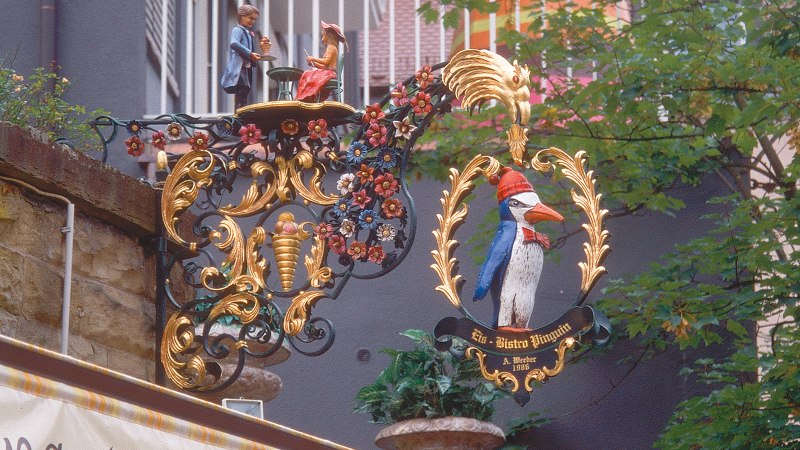 An artistically decorated sign with a penguin and figures, surrounded by floral ornaments, hangs on a wall on Eugensplatz., © Stuttgart-Marketing GmbH An artistically decorated sign with a penguin and figures, surrounded by floral ornaments, hangs on a wall on Eugensplatz., © Stuttgart-Marketing GmbH
