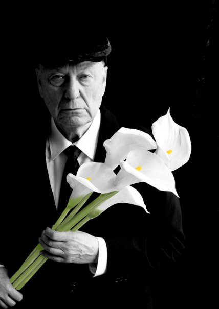An older man in a suit and cap holds a bouquet of white calla lilies against a black background. The expression is serious and thoughtful., © Theaterhaus Stuttgart e.V. An older man in a suit and cap holds a bouquet of white calla lilies against a black background. The expression is serious and thoughtful., © Theaterhaus Stuttgart e.V.