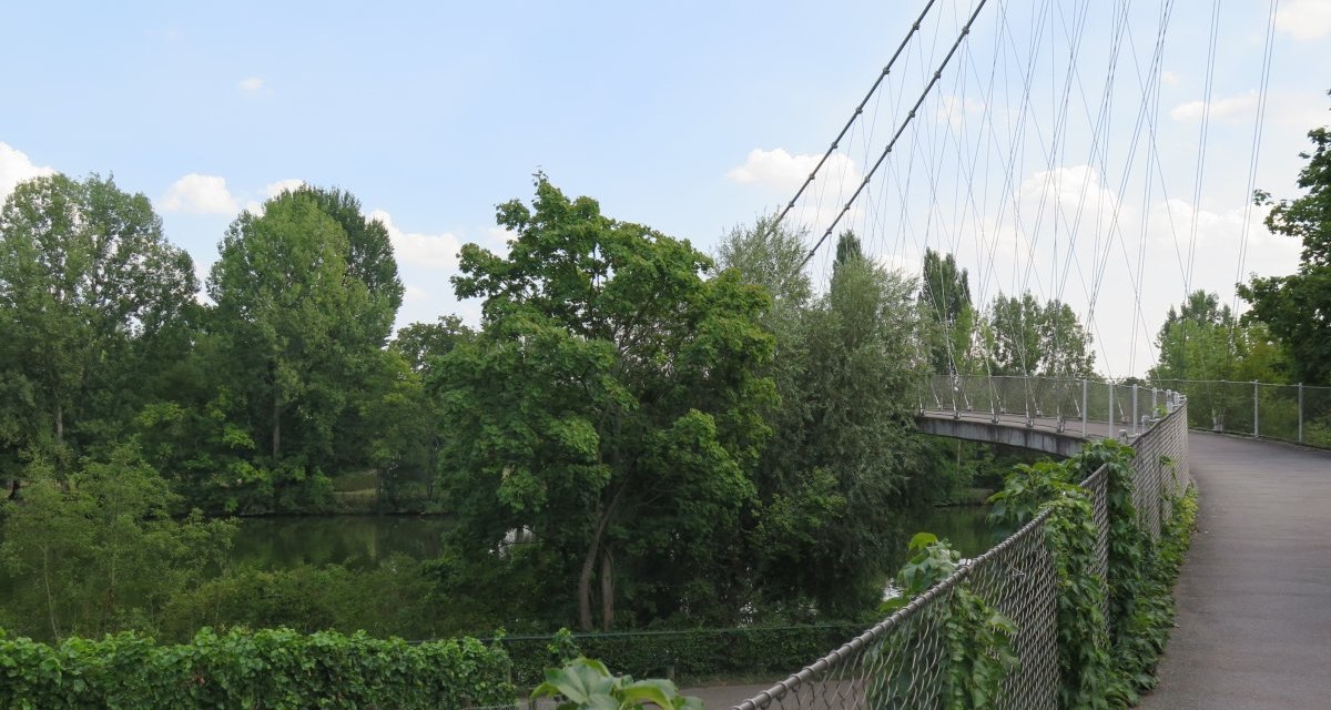 The Max-Eyth-Steg is a pedestrian bridge that crosses a river, surrounded by lush greenery and trees. The sky is slightly cloudy., © Stuttgart-Marketing GmbH The Max-Eyth-Steg is a pedestrian bridge that crosses a river, surrounded by lush greenery and trees. The sky is slightly cloudy., © Stuttgart-Marketing GmbH