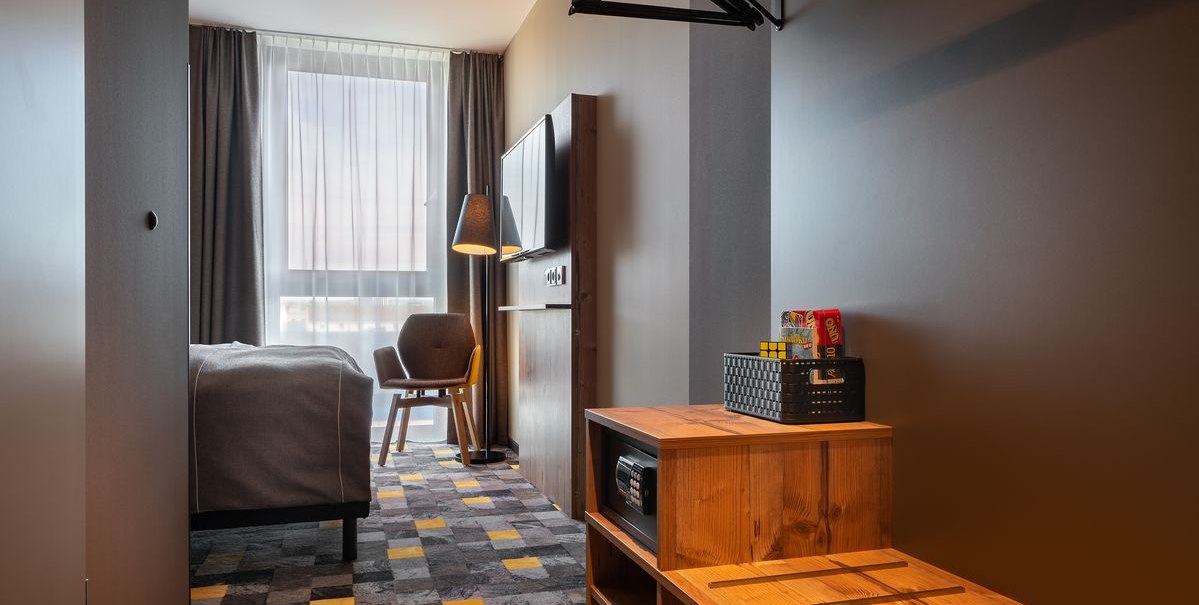 Modernes Hotelzimmer mit grauem Teppich, Bett, Sessel, Fernseher und Holzmöbeln. Ein Fenster mit Vorhängen lässt Tageslicht herein., © Novum Hospitality