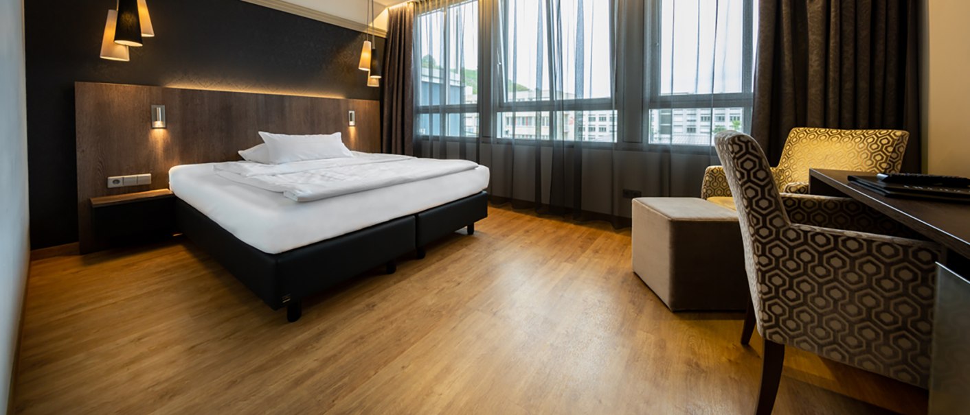 Modernes Hotelzimmer mit großem Bett, Holzboden, stilvollen Lampen und gemütlichen Sesseln vor großen Fenstern., © Kronenhotel Stuttgart