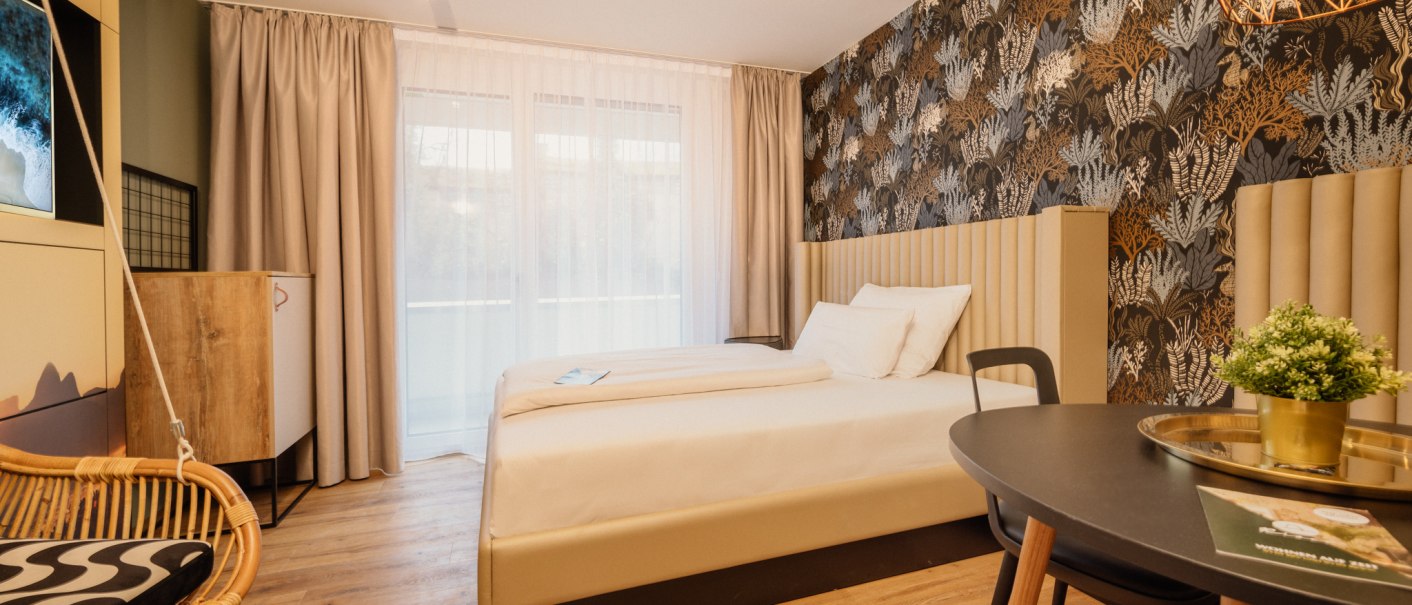 Modern apartment room with double bed, stylish wallpaper, hanging chair and table with plant., © Rioca Stuttgart Posto 4 GmbH Modern apartment room with double bed, stylish wallpaper, hanging chair and table with plant., © Rioca Stuttgart Posto 4 GmbH