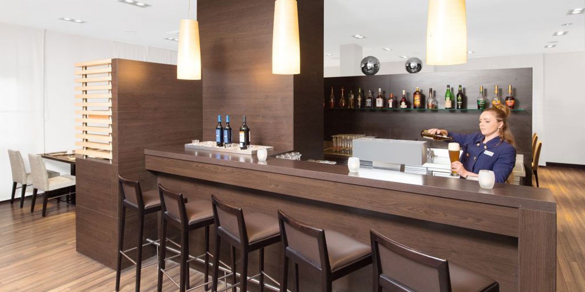 Moderne Bar mit Holztheke, Barhockern und einer Frau, die ein Getränk einschenkt. Im Hintergrund sind Flaschen auf Regalen zu sehen., © NH Stuttgart Sindelfingen Moderne Bar mit Holztheke, Barhockern und einer Frau, die ein Getränk einschenkt. Im Hintergrund sind Flaschen auf Regalen zu sehen., © NH Stuttgart Sindelfingen