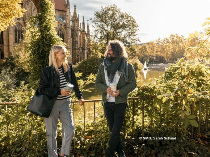 Zwei Personen spazieren lächelnd mit Kaffeebechern vor einer Kirche in Stuttgart. Im Hintergrund ein Teich mit Springbrunnen und herbstlicher Vegetation., © Stuttgart Marketing GmbH Zwei Personen spazieren lächelnd mit Kaffeebechern vor einer Kirche in Stuttgart. Im Hintergrund ein Teich mit Springbrunnen und herbstlicher Vegetation., © Stuttgart Marketing GmbH