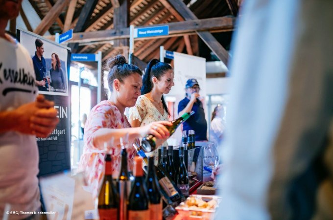 Two women are pouring wine at an event. Advertising banners and wine bottles can be seen in the background., © Stuttgart Marketing GmbH Two women are pouring wine at an event. Advertising banners and wine bottles can be seen in the background., © Stuttgart Marketing GmbH