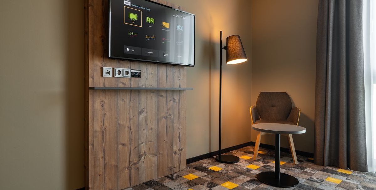 Modernes Hotelzimmer mit Holzverkleidung, Flachbildfernseher, Stehlampe, Sessel und kleinem Tisch auf gemustertem Teppichboden., © Novum Hospitality