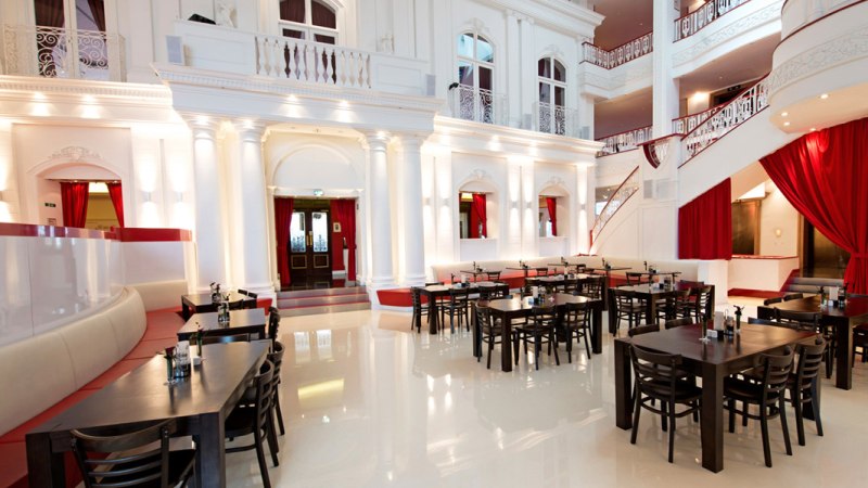Elegantes Restaurant mit hohen wei&szlig;en W&auml;nden, roten Vorh&auml;ngen und dunklen Holztischen. Die Architektur ist klassisch mit S&auml;ulen und Balkonen., &copy; Dormero Hotel Stuttgart