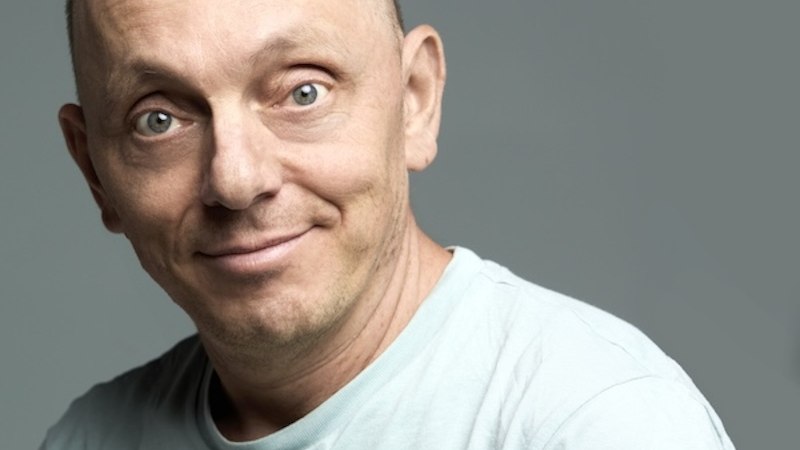 A bald man in a light-colored T-shirt smiles at the camera. The background is gray., © Renitenztheater Stuttgart e.V. A bald man in a light-colored T-shirt smiles at the camera. The background is gray., © Renitenztheater Stuttgart e.V.