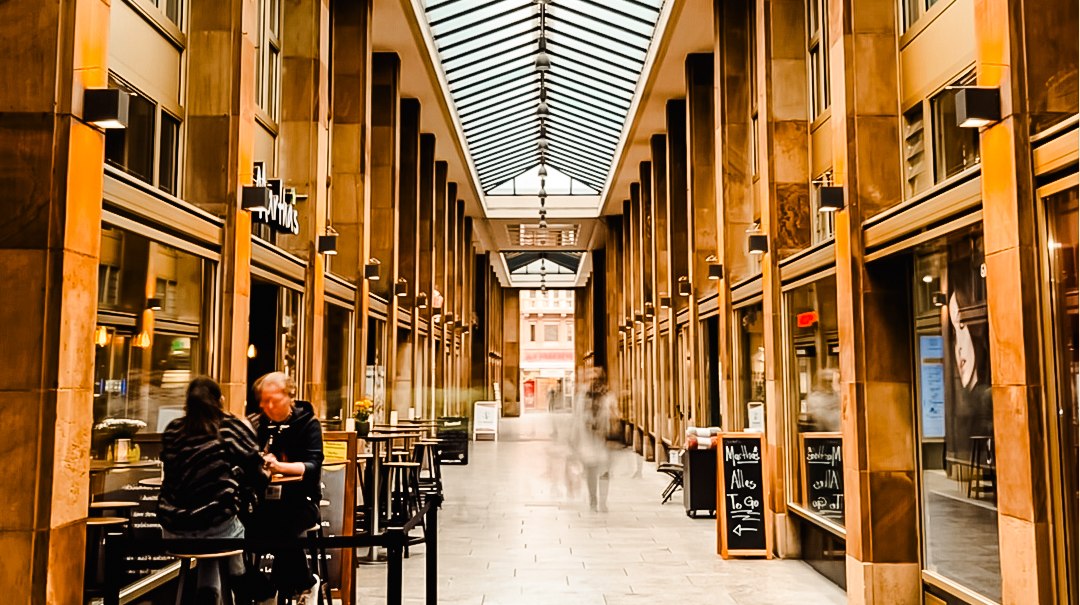 Eine überdachte Passage mit Glasdach, flankiert von Geschäften. Menschen sitzen an einem Café, während andere durch die Passage gehen., © Königsbau Eine überdachte Passage mit Glasdach, flankiert von Geschäften. Menschen sitzen an einem Café, während andere durch die Passage gehen., © Königsbau