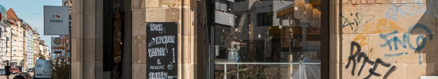 Café mit Außenbestuhlung, Graffiti an der Wand, Lichterketten über dem Eingang. Schild mit "Kaffee & Kuchen". Stadtstraße im Hintergrund., © SMG, Sarah Schmid Café mit Außenbestuhlung, Graffiti an der Wand, Lichterketten über dem Eingang. Schild mit "Kaffee & Kuchen". Stadtstraße im Hintergrund., © SMG, Sarah Schmid