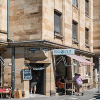 Cafébar Auszeit on a street corner with a red awning. People are sitting outside, street signs and cars are visible., © SMG, Sarah Schmid Cafébar Auszeit on a street corner with a red awning. People are sitting outside, street signs and cars are visible., © SMG, Sarah Schmid
