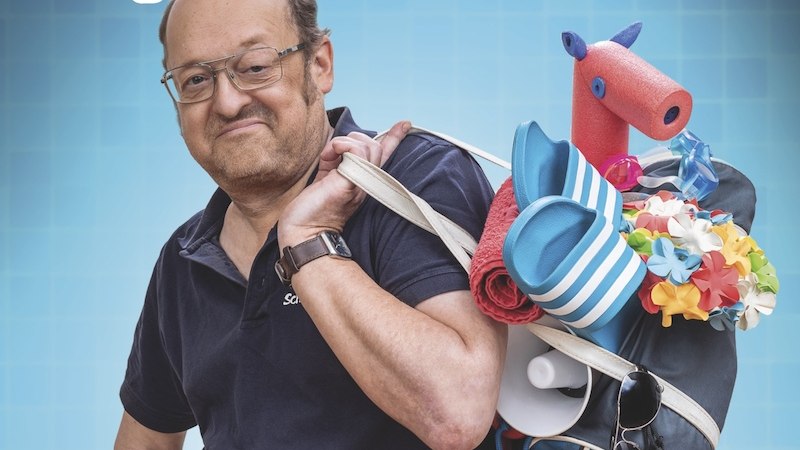 A man with glasses carries a bag full of swimming gear, including flippers and a pool noodle horse, in front of a blue background with the words 'Spassbad'., &copy; Renitenztheater Stuttgart e.V.