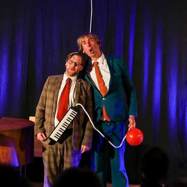 Zwei M&auml;nner in Anz&uuml;gen stehen auf einer B&uuml;hne. Einer h&auml;lt eine Melodica, der andere einen roten Ballon. Sie wirken humorvoll und unterhaltsam., &copy; Theaterhaus Stuttgart e.V.