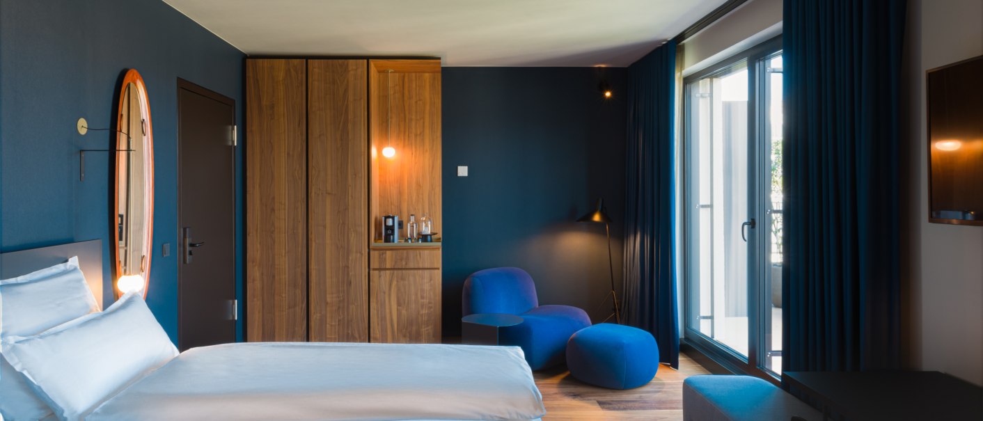Moderne Hotelsuite mit blauen Wänden, Holzmöbeln und einem großen Fenster. Ein Doppelbett, blaue Sessel und ein runder Spiegel sind sichtbar., © EmiLu Hotel GmbH
