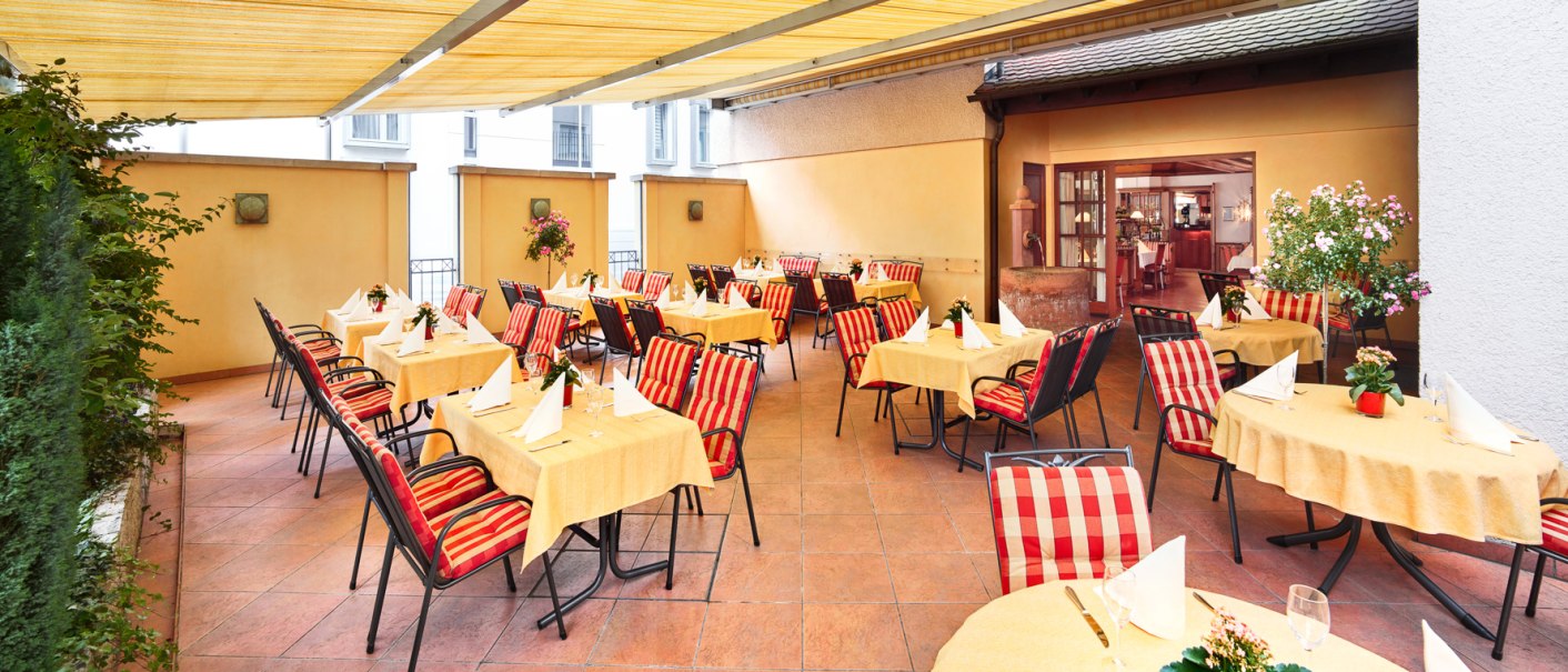 Terrasse eines Restaurants mit gelben Tischdecken, gestreiften Stühlen und Pflanzen. Überdachung bietet Schatten. Eingang zum Innenbereich sichtbar., © Hirsch Hotel