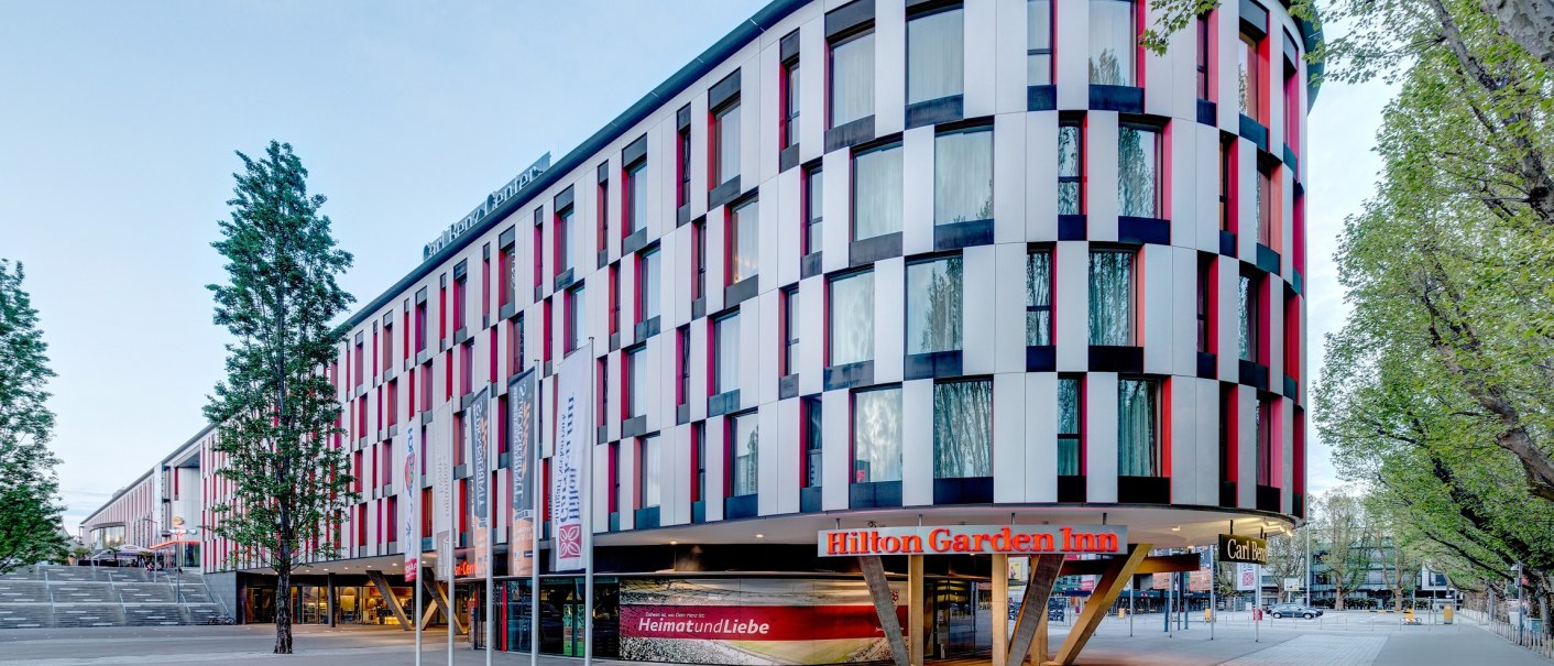 Modernes Hotelgebäude mit bunter Fassade, Hilton Garden Inn Stuttgart Neckarpark. Vor dem Gebäude stehen Fahnenmasten, umgeben von Bäumen., © Hilton Garden Inn Stuttgart Neckarpark