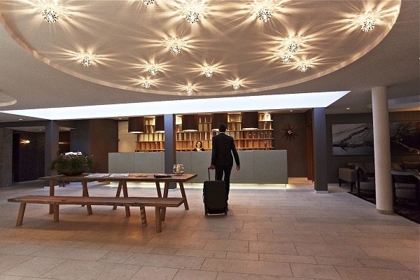 Ein Mann mit Koffer steht an der Rezeption des Waldhotels Stuttgart. Die Decke ist mit stilvollen Leuchten dekoriert., © Waldhotel Stuttgart GmbH