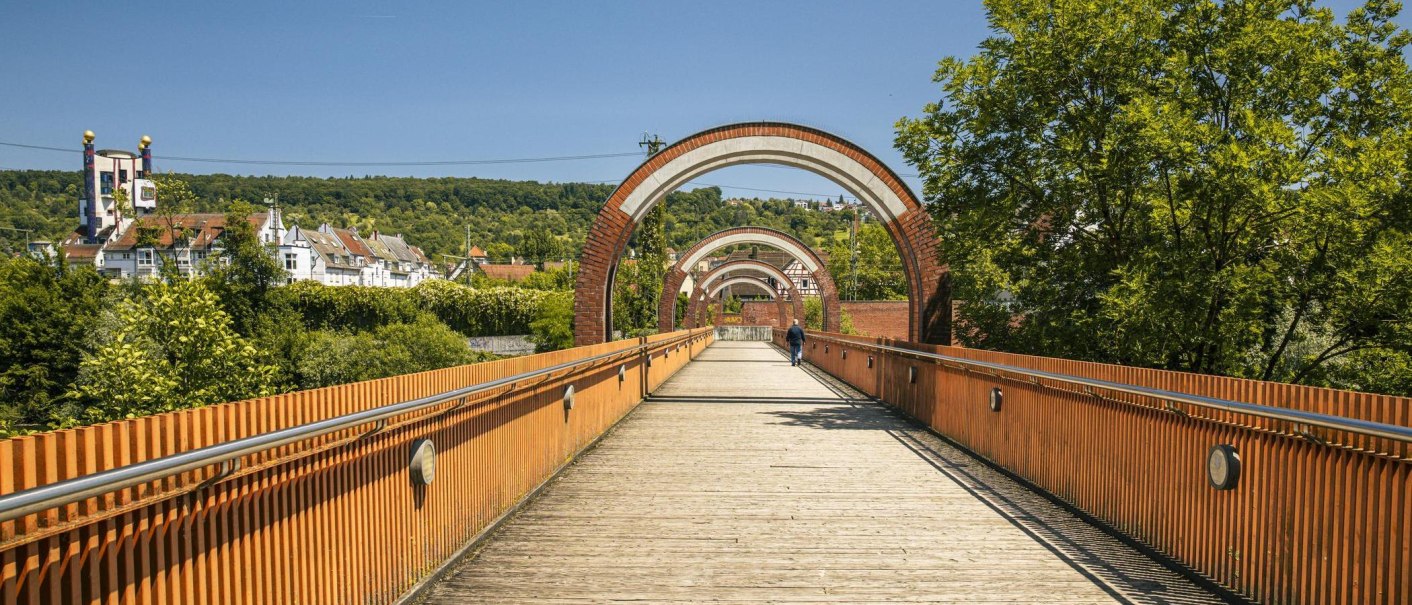 A wooden footbridge in Plochingen, featuring several arches, winds through a green landscape. Buildings and trees can be seen in the background., © SMG Stuttgart Marketing GmbH - Sarah Schmid A wooden footbridge in Plochingen, featuring several arches, winds through a green landscape. Buildings and trees can be seen in the background., © SMG Stuttgart Marketing GmbH - Sarah Schmid