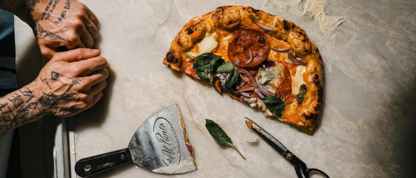 Halb gegessene Pizza mit Basilikum und Zwiebeln auf hellem Stein, daneben Pizzaschneider und Schere; links zwei tätowierte Hände., © Connective Media Halb gegessene Pizza mit Basilikum und Zwiebeln auf hellem Stein, daneben Pizzaschneider und Schere; links zwei tätowierte Hände., © Connective Media