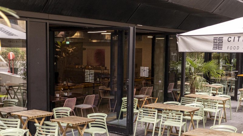 Außenbereich eines Cafés mit Holztischen und grünen Stühlen. Eine Markise trägt den Schriftzug 'Citizen Long'. Pflanzen dekorieren den Bereich., © SMG, Sarah Schmid