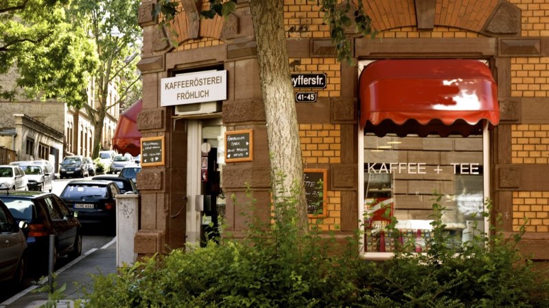 Entrance to the Fröhlich coffee roastery on a street corner with a red awning and signs. Cars are parked along the street, trees provide shade., © Kaffeerösterei Fröhlich Entrance to the Fröhlich coffee roastery on a street corner with a red awning and signs. Cars are parked along the street, trees provide shade., © Kaffeerösterei Fröhlich
