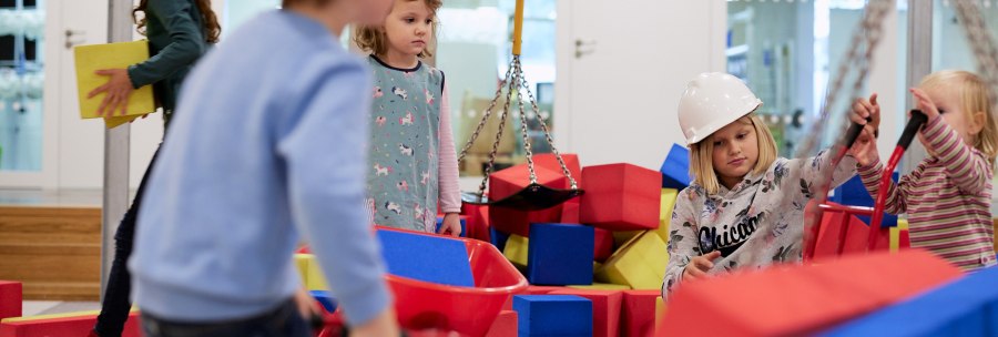 Kinder mit Bauhelmen spielen auf einer Indoor-Baustelle mit bunten Bauklötzen und Schubkarren., © Julia Ochs Kinder mit Bauhelmen spielen auf einer Indoor-Baustelle mit bunten Bauklötzen und Schubkarren., © Julia Ochs