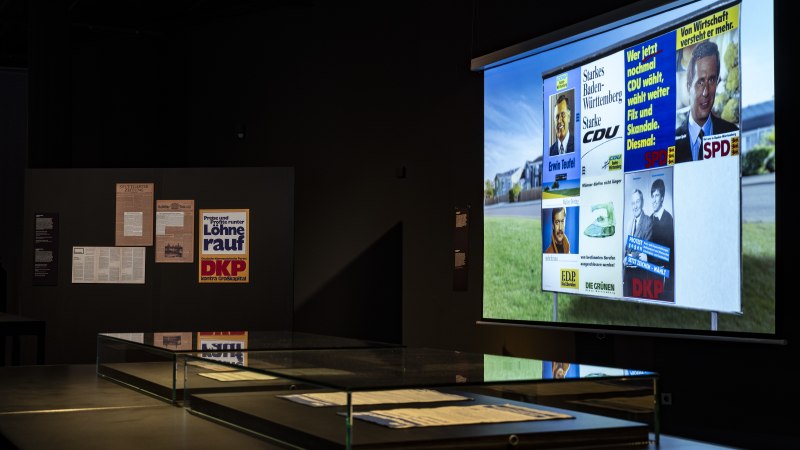 Ausstellung im Haus der Geschichte Baden-Württemberg zeigt historische Wahlplakate und Dokumente. Dunkler Raum mit beleuchteten Exponaten., © HdGBW Ausstellung im Haus der Geschichte Baden-Württemberg zeigt historische Wahlplakate und Dokumente. Dunkler Raum mit beleuchteten Exponaten., © HdGBW
