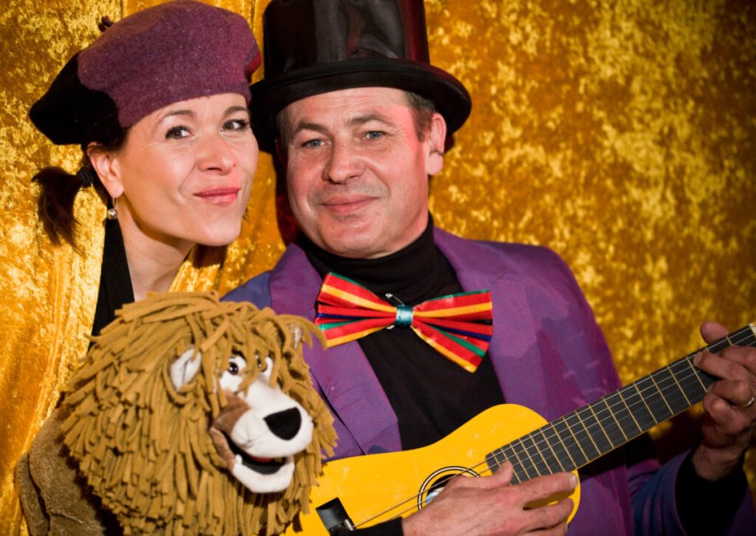 Zwei Personen in bunten Kostümen, eine mit Löwenpuppe, die andere mit Gitarre, vor goldenem Hintergrund. Beide lächeln in die Kamera., © Liedertheater Altmann